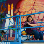 Yreina D. Cervántez and La Ofrenda mural woman on scaffold in front of mural