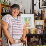 Yreina D. Cervántez....photo by Lluvia Higuera woman standing and smiling amid artwork